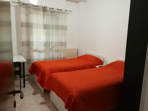 A cozy hotel room features two beds with vibrant red bedding, a small desk by the window with sheer curtains, evoking a simple and functional atmosphere ideal for budget travelers seeking comfort.
