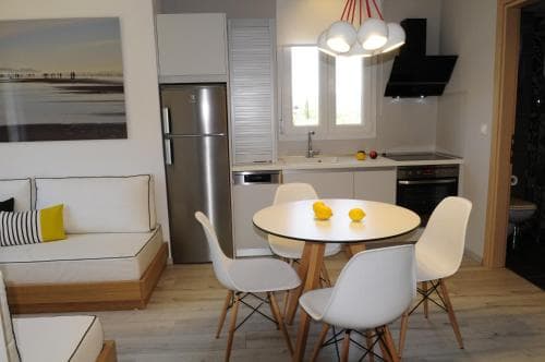 A cozy modern apartment kitchen features a round dining table with lemons, white chairs, stainless steel appliances, and a bright window, ideal for travelers seeking a comfortable self-catering stay.