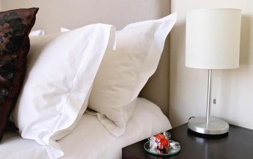 A cozy hotel room corner features plush white pillows on a bed beside a sleek bedside table with a modern lamp and a small plate of candies, ideal for travelers seeking comfort and relaxation.