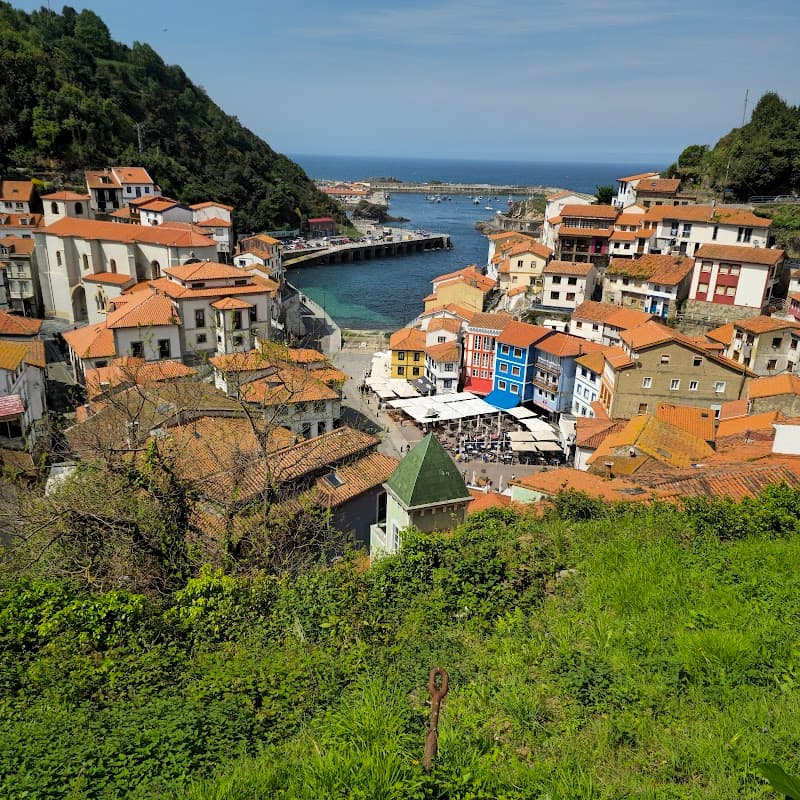 A picturesque coastal village with vibrant orange-roofed houses nestled among lush greenery, overlooking a serene harbor, ideal for travelers seeking a charming seaside retreat in Asturias, Spain.