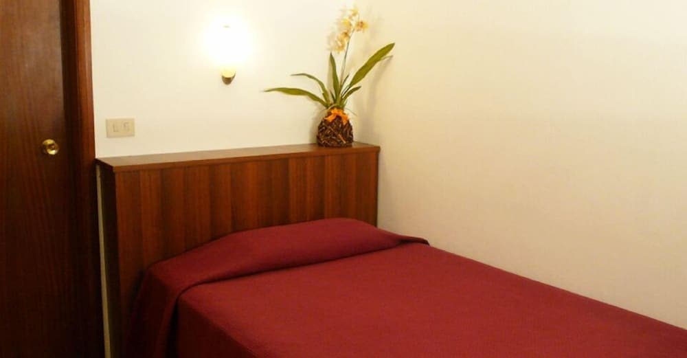A cozy hotel room features a neatly made bed with a rich red cover, a wooden headboard adorned with a pineapple vase holding flowers, and soft lighting, ideal for travelers seeking comfort and simplicity.