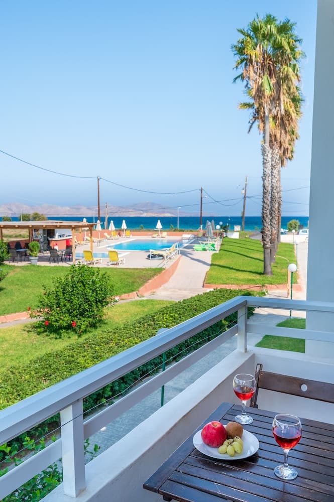 A serene balcony view showcases a tranquil poolside area with lounge chairs, a towering palm tree, and a picturesque sea backdrop, perfect for a relaxing vacation retreat.