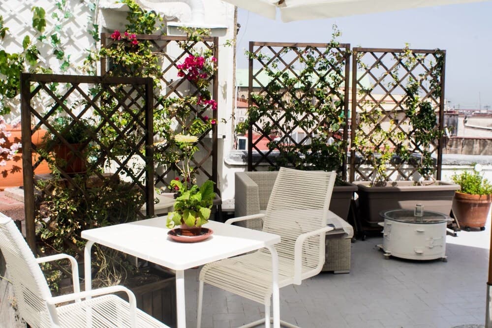 A serene rooftop terrace adorned with lush greenery and vibrant flowers offers a cozy spot for relaxation, featuring white wicker furniture and a small table perfect for intimate gatherings or enjoying the cityscape view.