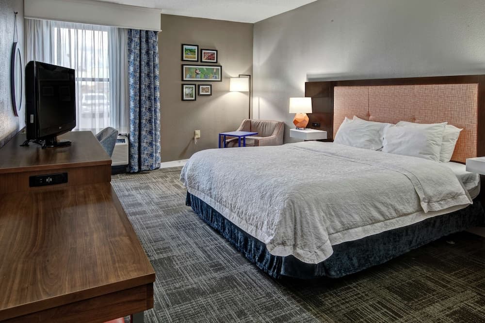 A cozy hotel room features a neatly made bed with plush pillows, a wooden desk, a TV, and a window with patterned curtains, creating a comfortable and inviting atmosphere ideal for travelers seeking relaxation.