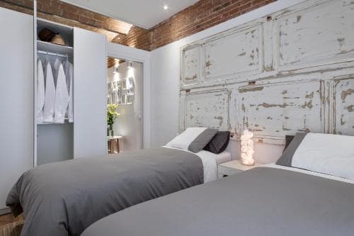 A cozy urban loft bedroom features twin beds with gray linens, rustic white-paneled walls, exposed brick accents, and a walk-in closet, ideal for travelers seeking a charming and comfortable stay in a city retreat.