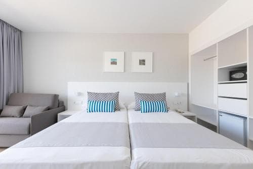 A cozy hotel room features twin beds with blue-striped pillows, a modern sofa, and minimalist decor, ideal for travelers seeking comfort and simplicity in a clean, contemporary setting.