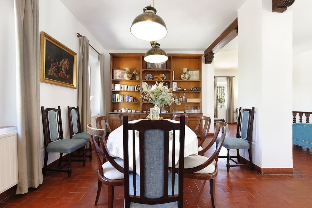 A cozy dining room with terracotta floors, wooden chairs around a round table set with a floral centerpiece, a built-in bookshelf filled with books and decor, and warm lighting creates an inviting atmosphere ideal for family gatherings or intimate dinners.