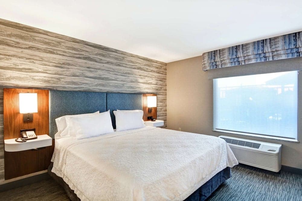 A cozy hotel room features a neatly made bed with white linens, modern bedside tables with lamps, a textured wall accent, and a large window with natural light, ideal for travelers seeking comfort and relaxation.