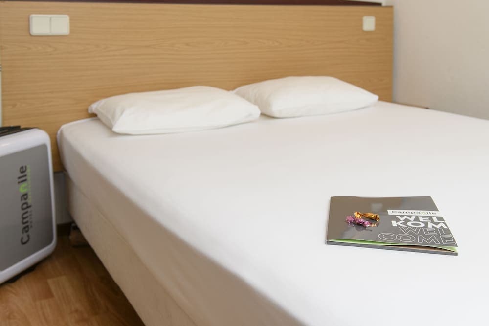 A cozy hotel room features a neatly made bed with white linens, a wooden headboard, and a welcoming brochure with chocolates, ideal for travelers seeking comfort and simplicity.