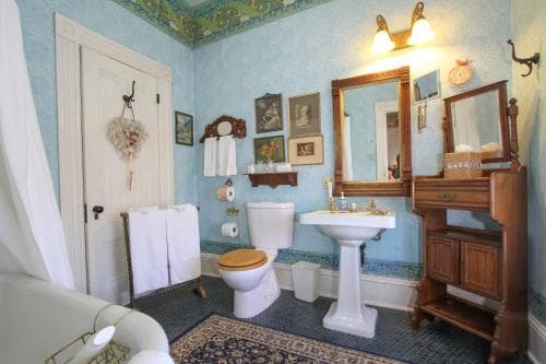 A charming vintage bathroom with light blue walls, ornate mirrors, wooden furnishings, and a patterned rug exudes a cozy, nostalgic atmosphere perfect for a heritage hotel stay.