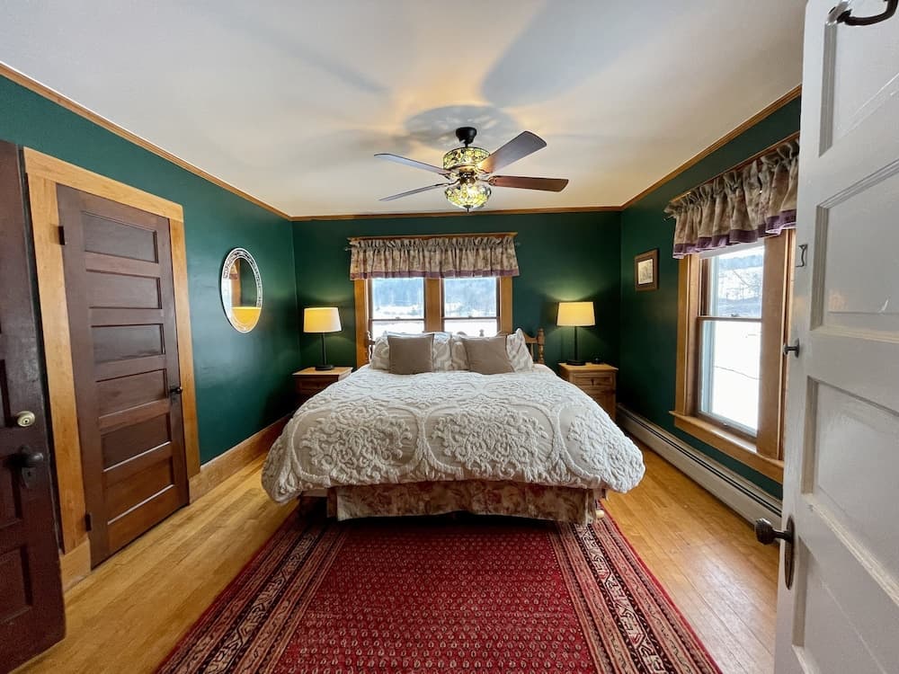 A cozy bedroom with green walls, wooden floors, and a red patterned rug exudes warmth, featuring a bed with white bedding, two bedside lamps, a ceiling fan with a stained glass light, and large windows offering a serene view, ideal for travelers seeking a tranquil retreat.
