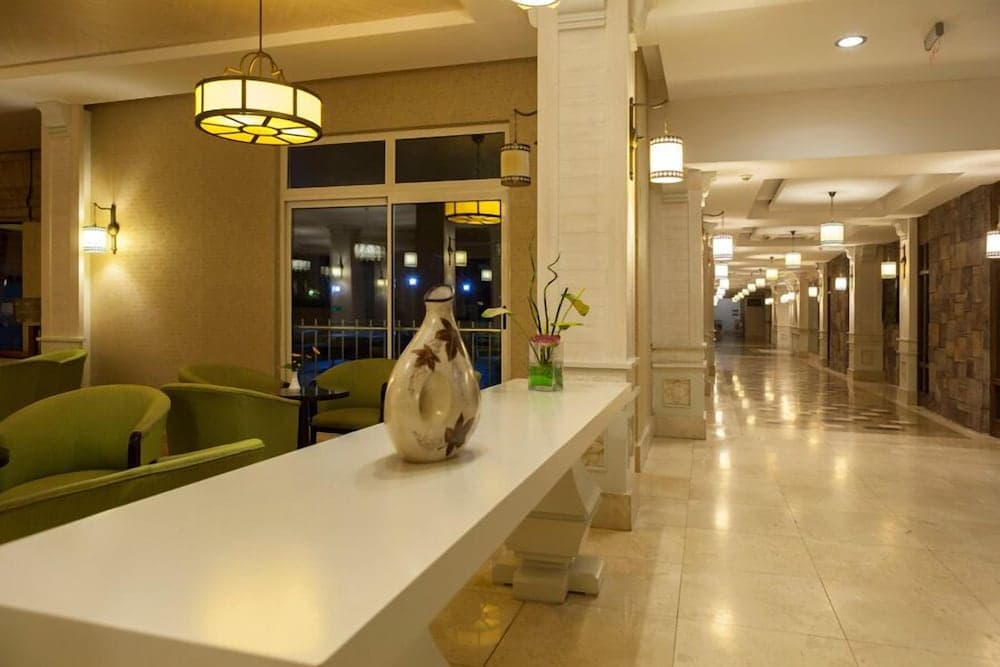 A warmly lit hotel lobby features elegant green armchairs, a sleek white table adorned with decorative vases, and a long corridor lined with soft lighting, creating an inviting atmosphere ideal for travelers seeking comfort and style.