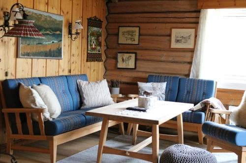 A cozy cabin interior features wooden walls adorned with framed artwork, blue cushioned seating, and a central table, creating a warm and inviting atmosphere ideal for a relaxing getaway.