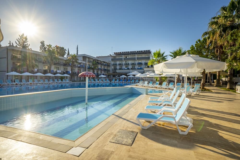 A serene hotel poolside scene bathed in sunlight features lounge chairs under white umbrellas, surrounded by palm trees and modern buildings, ideal for travelers seeking relaxation and leisure.