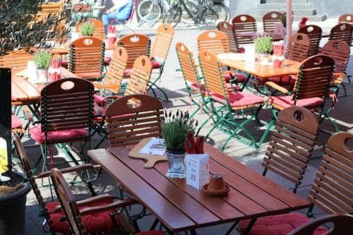 A cozy outdoor café scene with wooden tables, red-cushioned chairs, and potted plants creates an inviting atmosphere perfect for travelers seeking a relaxing spot to enjoy coffee or a meal.
