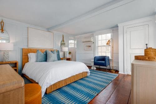 A cozy, well-lit hotel bedroom features a neatly made bed with blue accents, wooden floors, and a vibrant rug, creating a serene and inviting atmosphere ideal for travelers seeking comfort and relaxation.