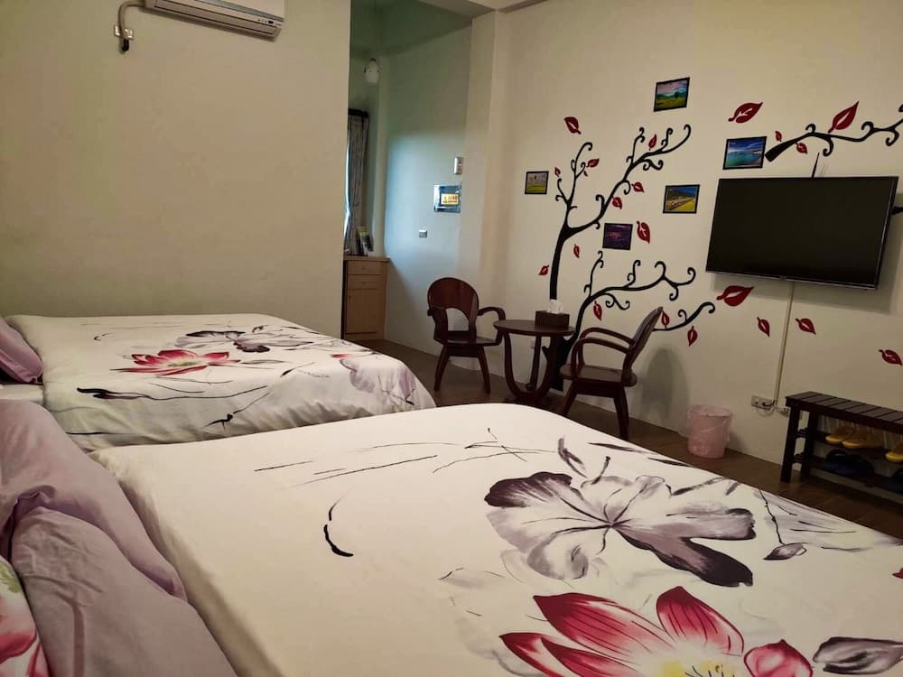 A cozy hotel room features two beds with floral bedding, a wall-mounted TV, decorative tree art, and a small seating area, ideal for travelers seeking comfort and relaxation.
