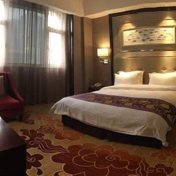 A cozy hotel room features a neatly made bed with plush pillows, warm lighting, patterned carpeting, and a red armchair, creating an inviting atmosphere ideal for travelers seeking comfort and relaxation.