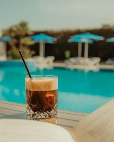 A refreshing iced coffee sits on a table by a serene poolside, surrounded by lounge chairs and blue umbrellas under a sunny sky, perfect for a relaxing vacation getaway.