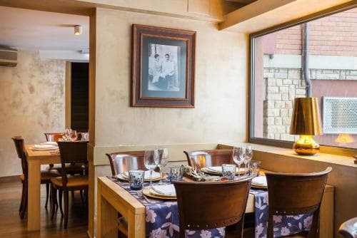 A cozy restaurant interior features wooden tables set for dining, framed artwork on the walls, and a warm ambiance enhanced by a golden lamp near a window, ideal for travelers seeking an intimate dining experience.