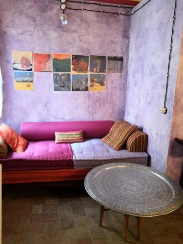A cozy, bohemian-style living room with purple walls adorned with colorful artwork, a vibrant sofa, and a decorative metal table, perfect for travelers seeking a unique and artistic accommodation experience.