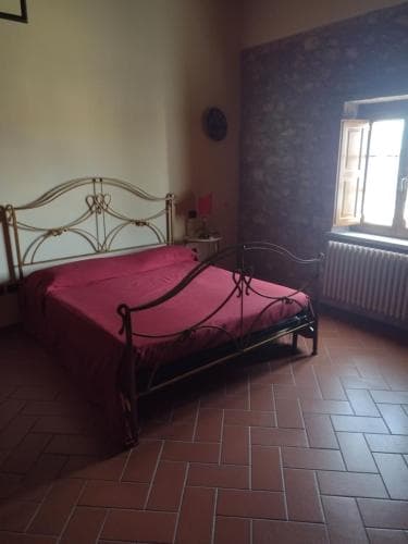 A cozy bedroom with a rustic charm features a metal bed adorned in red linens, terracotta tiled floors, and a bright window, ideal for travelers seeking a quaint and comfortable stay.