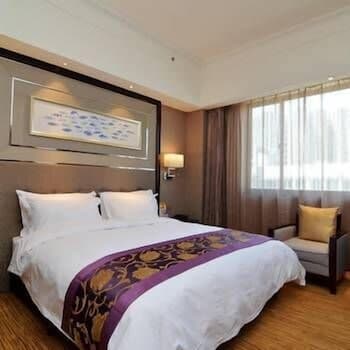 A cozy hotel room features a neatly made bed adorned with a purple and gold throw, framed artwork above the headboard, and a comfortable armchair by the window, ideal for travelers seeking relaxation and comfort.