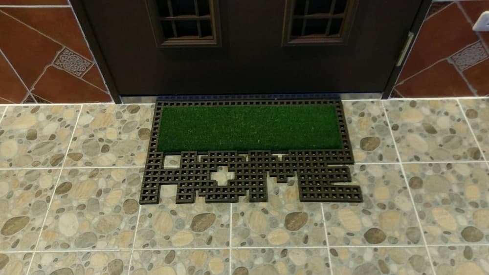A cozy home entrance features a pebble-tiled floor with a pixelated "HOME" doormat in front of a dark wooden door, ideal for travelers seeking a welcoming stay.
