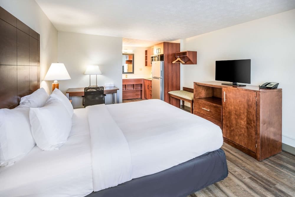 A cozy hotel room features a neatly made bed, wooden furnishings, a flat-screen TV, and an open kitchenette, creating a welcoming atmosphere ideal for travelers seeking comfort and convenience.