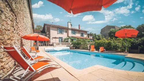 A serene outdoor poolside scene features vibrant orange umbrellas and loungers against a backdrop of rustic stone walls and charming houses, ideal for a relaxing vacation retreat.