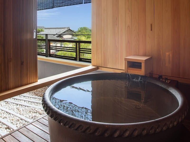 A serene indoor-outdoor onsen bath in Kyoto's Ukyo Ward offers a tranquil retreat with a wooden interior, stone tub, and a view of traditional rooftops and lush greenery, perfect for luxury travelers seeking relaxation.