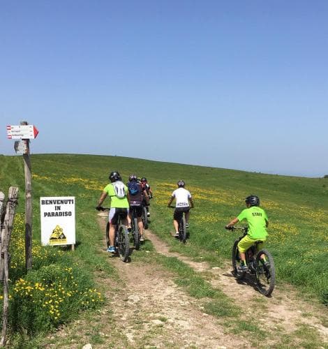 Cyclists pedal along a scenic dirt path through lush green hills under a clear blue sky, passing a welcoming sign that reads "Benvenuti in Paradiso," ideal for adventure seekers exploring serene countryside trails.