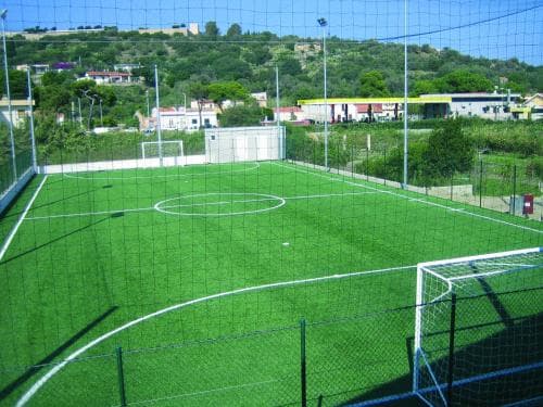 A vibrant artificial turf soccer field enclosed by netting offers an inviting spot for sports enthusiasts, set against a backdrop of lush green hills and quaint residential buildings, ideal for travelers seeking recreational activities in scenic locales.