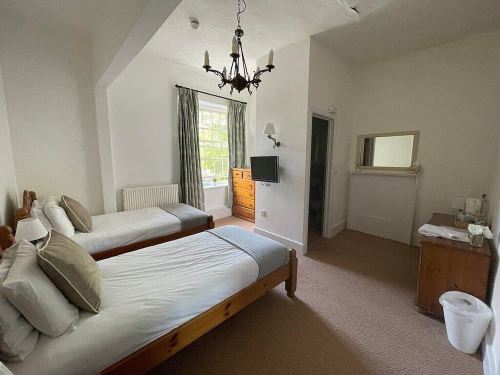 A cozy, well-lit hotel room features two neatly arranged single beds under a charming chandelier, with a window offering greenery views, ideal for travelers seeking comfort and tranquility.