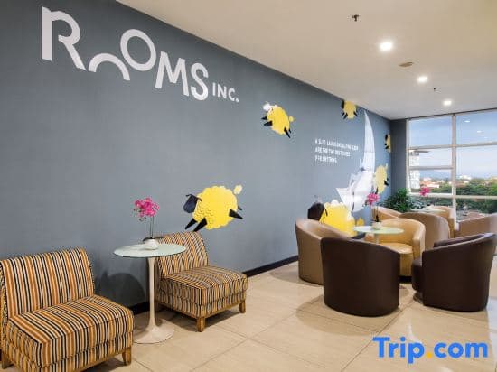 A cozy hotel lounge features vibrant striped chairs, modern tables, and playful yellow bird wall art under the "ROOMS INC." logo, creating an inviting atmosphere ideal for travelers seeking relaxation and comfort.