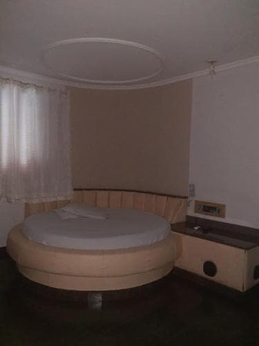 A cozy hotel room features a unique round bed with beige upholstery, soft curtains by the window, and a minimalist nightstand, ideal for travelers seeking a distinctive and comfortable stay.