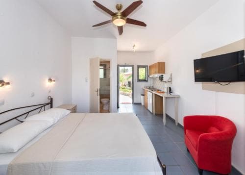 A cozy studio apartment features a neatly made bed, a vibrant red armchair, a compact kitchenette, and a ceiling fan, creating a welcoming atmosphere ideal for travelers seeking comfort and convenience in a compact living space.