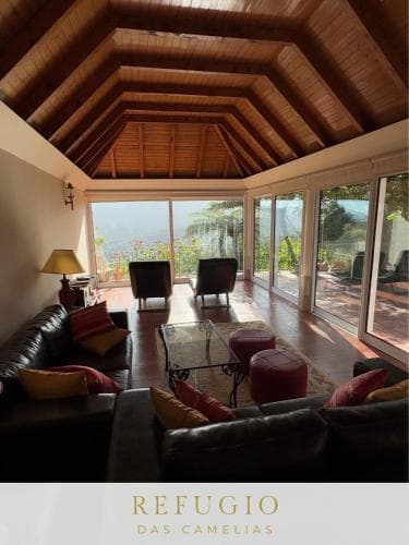 A cozy living room with leather sofas, vibrant cushions, and a wooden vaulted ceiling overlooks lush greenery through large glass doors, creating an inviting retreat ideal for relaxation and nature appreciation.