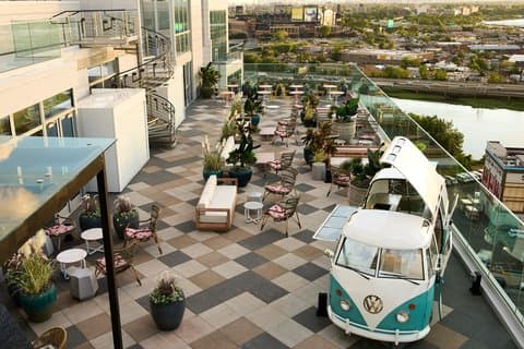 A vibrant rooftop terrace at the Renaissance New York Flushing Hotel features a cozy lounge area with potted plants, outdoor seating, and a vintage Volkswagen bus, offering a serene urban retreat with panoramic city views ideal for leisure travelers seeking relaxation.