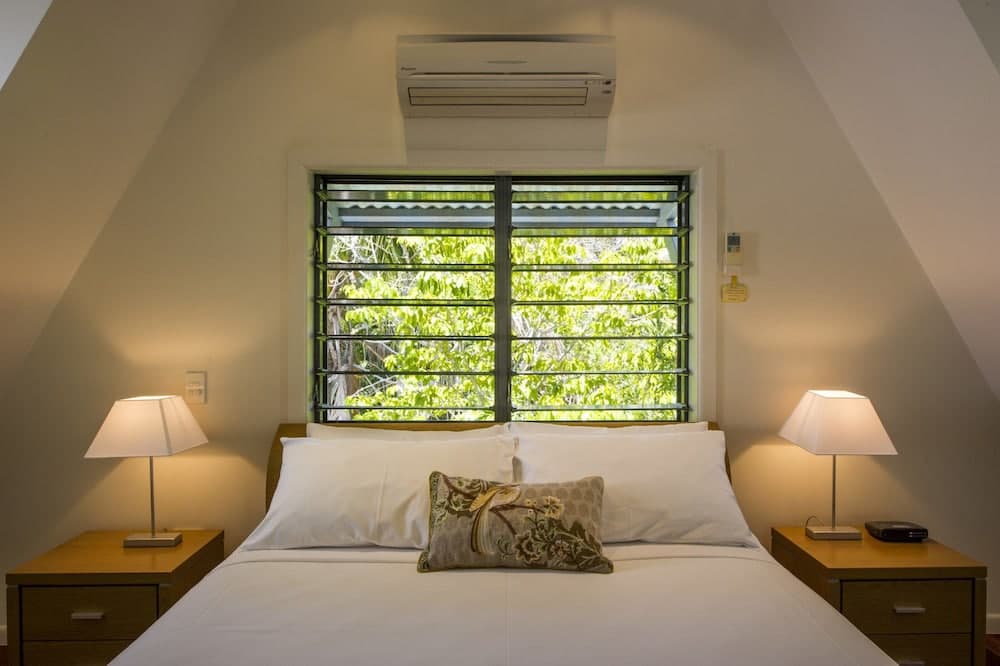 A cozy attic bedroom with sloped ceilings features a neatly made bed flanked by wooden nightstands with lamps, a window offering lush greenery views, and an air conditioner, ideal for travelers seeking a serene retreat.