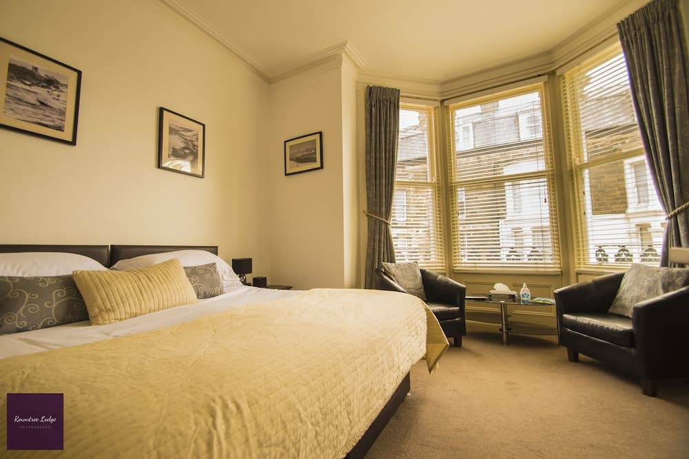 A cozy hotel room features a neatly made bed, plush armchairs by a bay window with city views, framed artwork on the walls, and a warm, inviting ambiance ideal for travelers seeking comfort and relaxation.