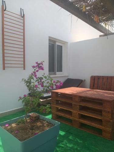 A cozy outdoor patio features a vibrant green floor, wooden pallet furniture, a blooming bougainvillea plant in a blue planter, and a climbing frame against white walls, creating a serene and inviting space perfect for relaxation or small gatherings.