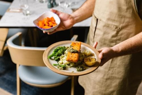 A chef presents a vibrant plate of tofu, broccoli, and potatoes in a modern dining setting, ideal for culinary enthusiasts exploring farm-to-table experiences.