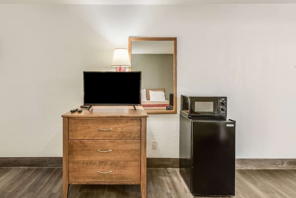 A cozy hotel room corner features a wooden dresser with a flat-screen TV, a mirror reflecting the bed, and a black mini-fridge with a microwave, ideal for travelers seeking comfort and convenience.