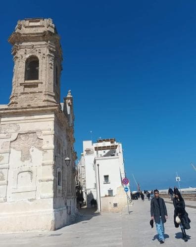 A sunny seaside promenade features historic architecture with a weathered bell tower, bustling pedestrians, and clear blue skies, ideal for cultural exploration and leisurely walks.