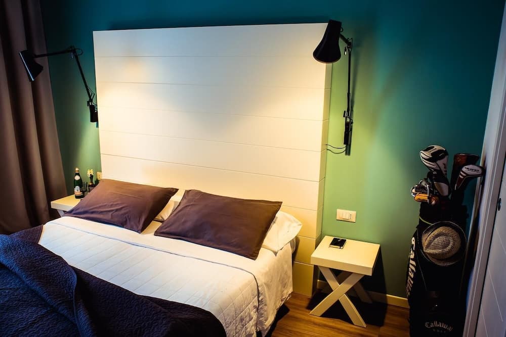 A cozy hotel room with a neatly made bed, teal walls, modern bedside lamps, and a golf bag by the door, ideal for travelers seeking comfort and convenience.