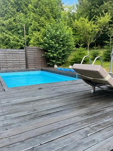 A serene backyard deck features a vibrant blue swimming pool surrounded by lush greenery, with a wicker lounge chair inviting relaxation, perfect for a tranquil getaway or family vacation.