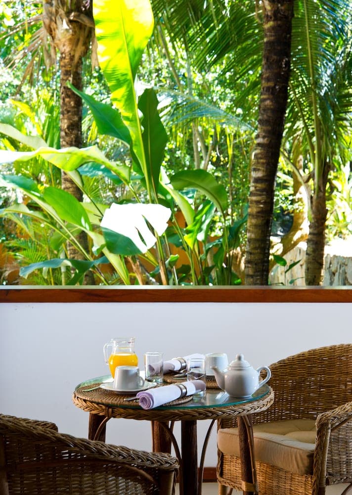 A serene tropical patio features wicker furniture set for breakfast with juice and tea, surrounded by lush greenery, ideal for a relaxing vacation getaway.