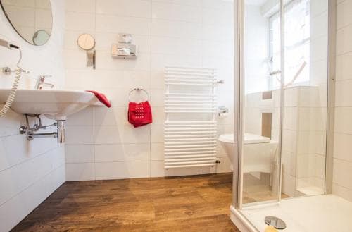 A modern bathroom with white tiled walls, wooden flooring, a glass shower enclosure, and a red towel adds a cozy touch, ideal for travelers seeking comfort in accommodations.