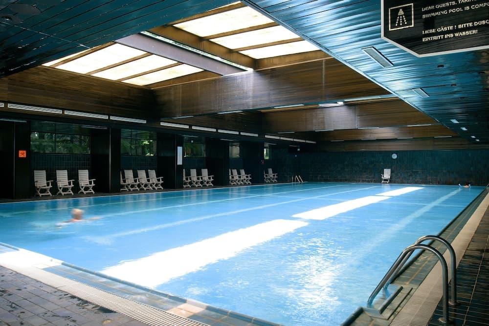 A serene indoor swimming pool with clear blue water, surrounded by lounge chairs under a modern glass ceiling, ideal for relaxation and leisure activities.